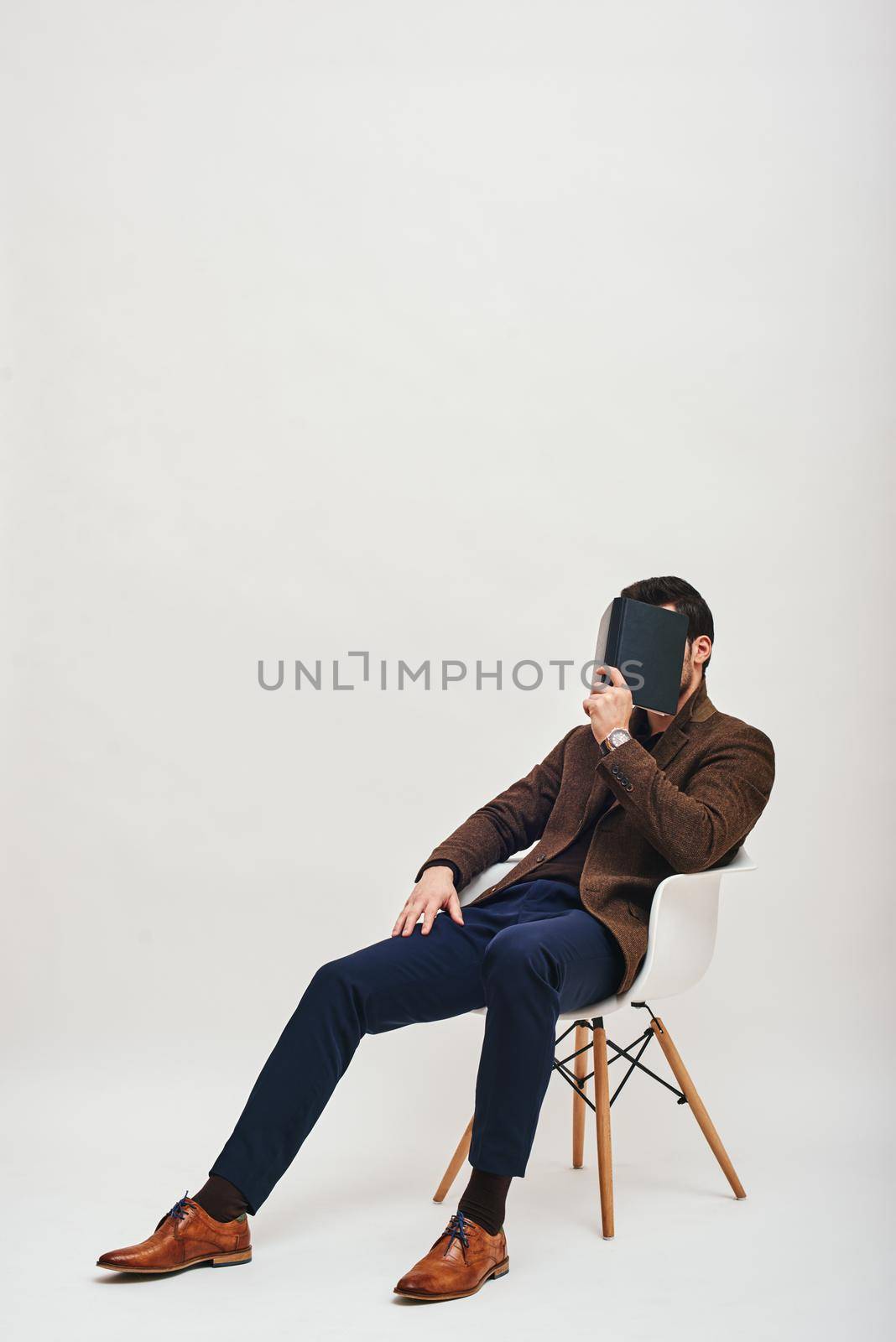 Full-length portrait of stylish man wearing blue trousers and brown jacket, sitting on a white chair and hiding his face behind a book