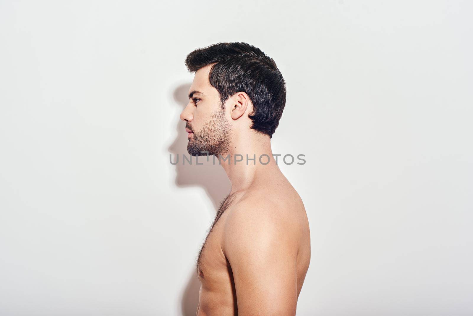 Portrait of young man with stylish hairstyle standing shirtless, isolated over white background. Side view