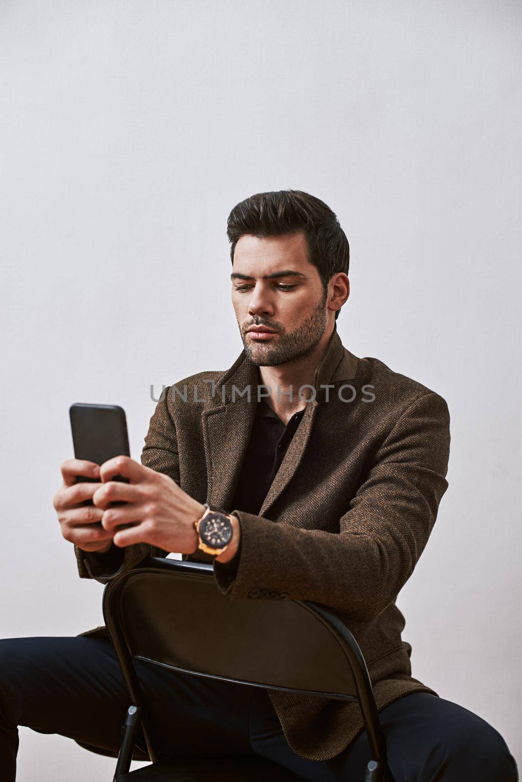 Isolated studio shot of handsome young male corporate worker with bristle and stylish haircut looking at his phone while sitting on a chair isolated over white background.