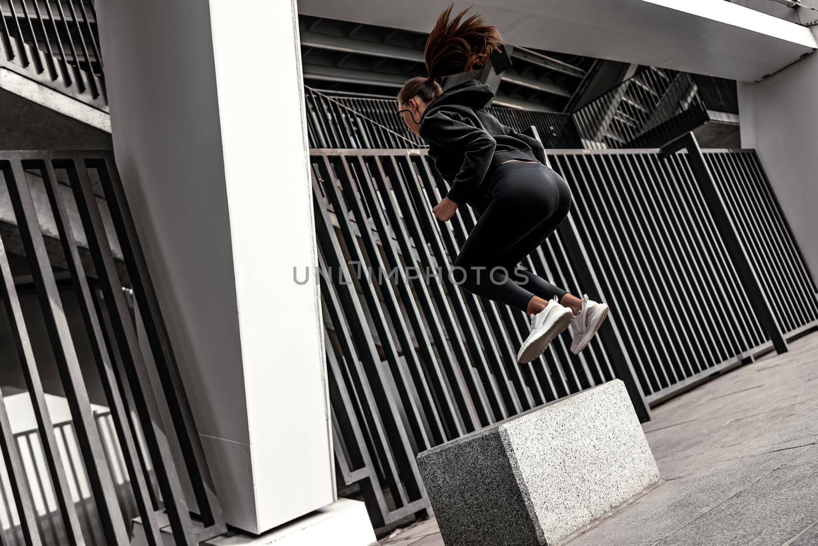 Young woman doing leg exercises at stoned step platform outside with city on background. Beautiful cauceuropean woman jumping over big step, squat exercise. Fitness and health concepts. Rear view