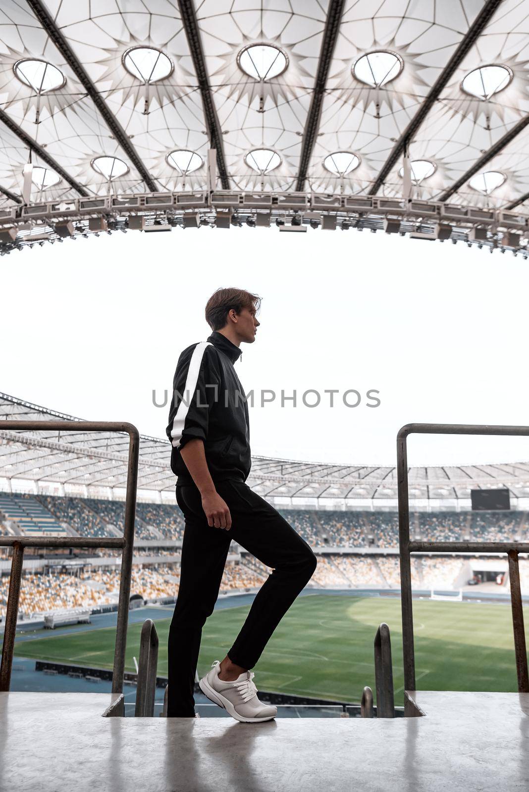 Young sportsman at big empty football stadium watching from the top. He looks to the stadium