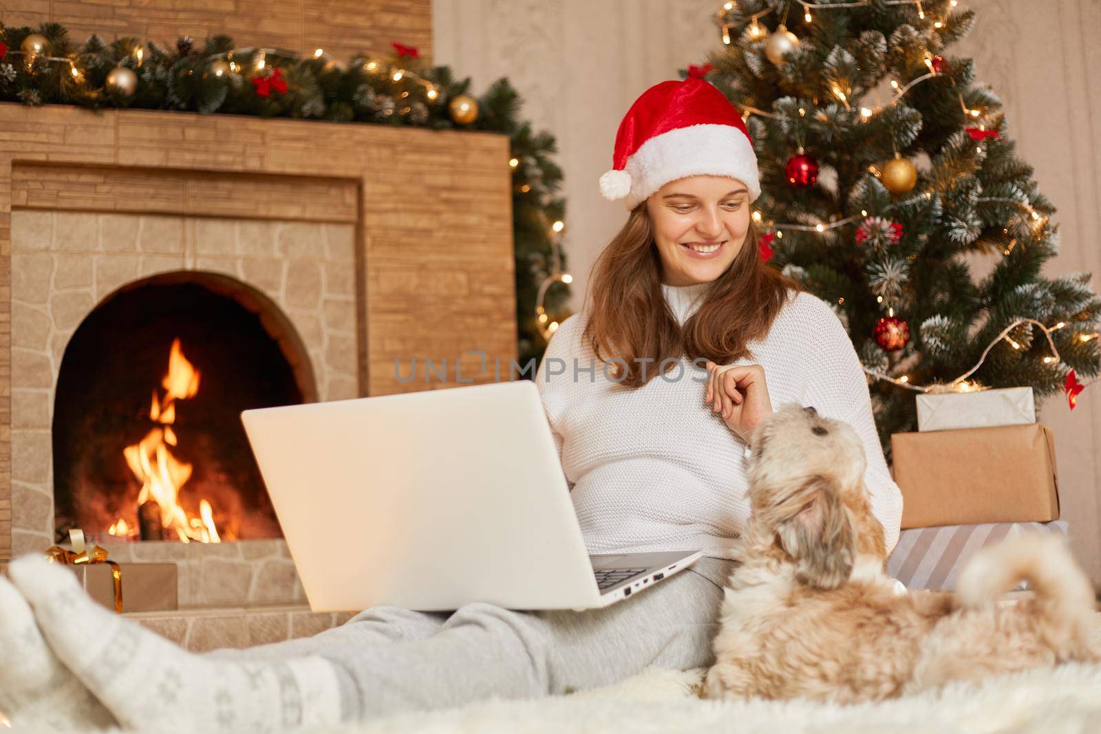 Charming woman sitting on floor with laptop, looks at her pet who sits near her, happy female wears red hat and casual sweater, lady with pekingese dog pose in festive Christmas room near fireplace.