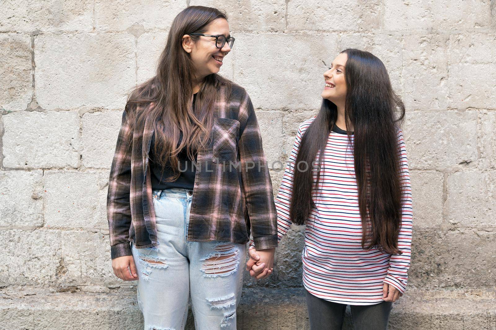 Two young lesbians holding each other and standing near a stone wall, smiling, happy. by HERRAEZ