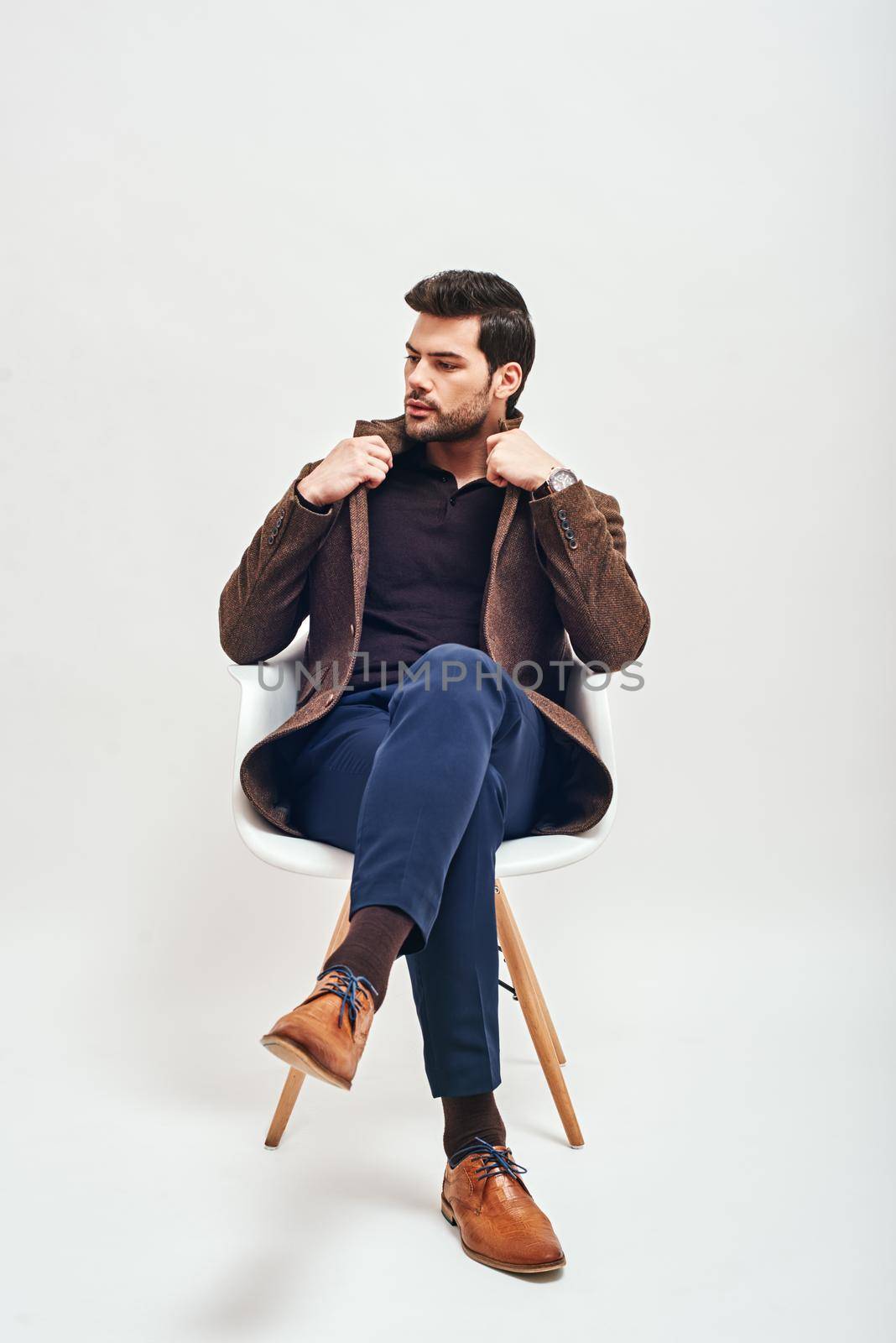 Full-length portrait of stylish dark-haired man wearing blue trousers and brown jacket, sitting on a white chair, crossing his legs and looking away isolated over white background