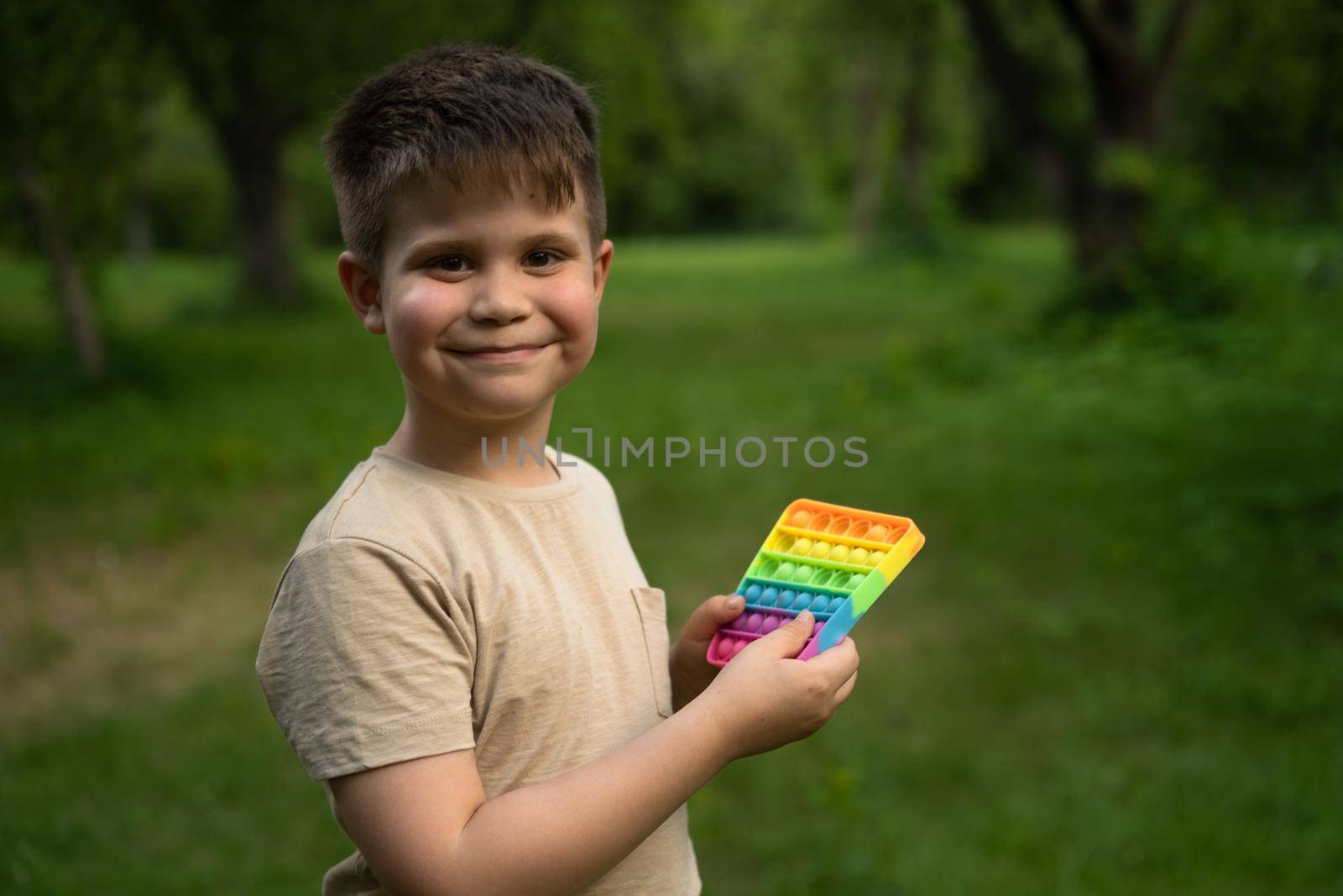 Pink-Cheeked Boy with Nice Dimples Clicks his New Pop It. Smiling 7 years guy having a walk in the park. Park Background. High quality photo