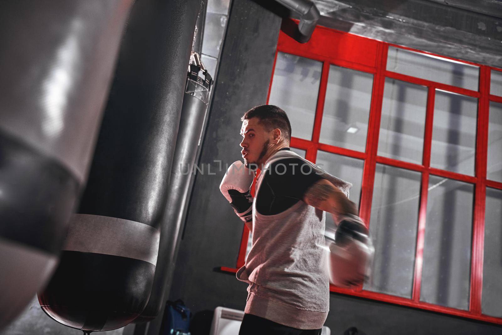 Handsome young muscular sportsman in sports clothing boxing on heavy punch bag. Hard training before big fight in colored boxing gym