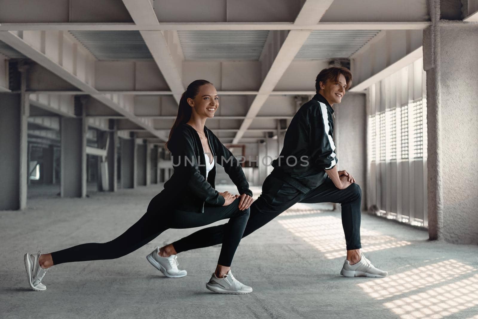 Young man and woman in sportive clothes are doing warming up exercises at parking near the stadium. Hands and legs stretching