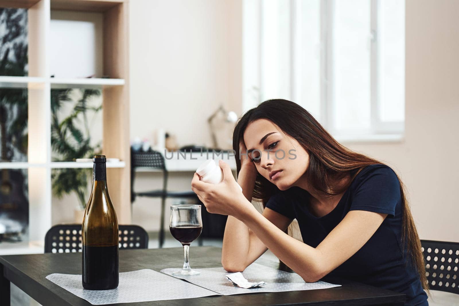Young dark-haired woman is going to drink a glass of red wine and take pills alone in kitchen at home. Female alcoholism concept. Protest in the treatment of alcohol addiction.