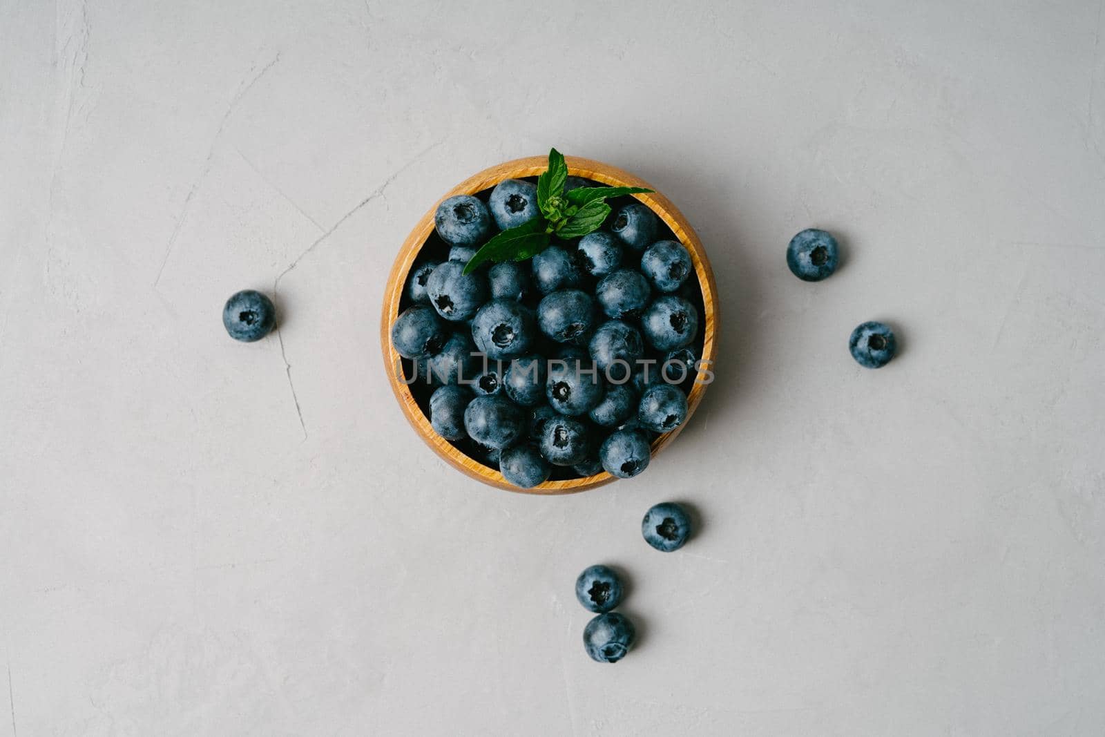 A small wooden bowl with berries on a gray background. Juicy and ripe blueberries in an eco-friendly dish. Top view of a bowl of blueberries. Berries on a gray textured background.