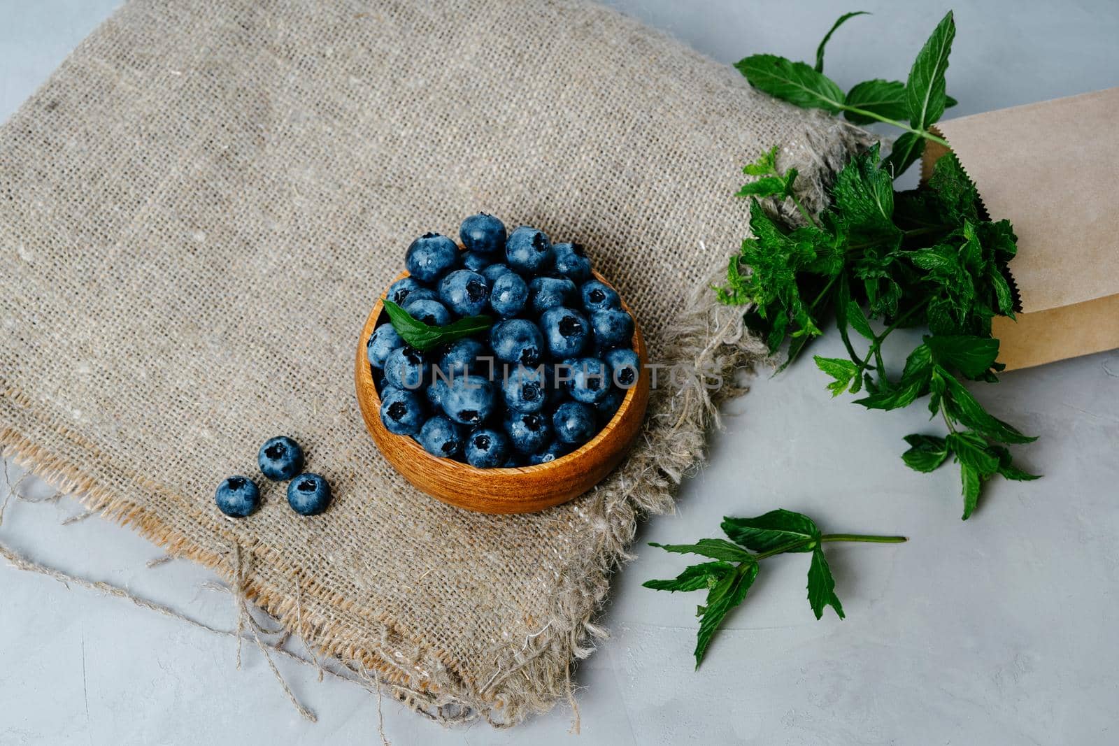 A small wooden bowl with berries. Juicy and ripe blueberries in an eco-friendly dish. Sprigs of fresh mint in a kraft bag. The concept of healthy eating and nutrition. Drops of water on blueberries.