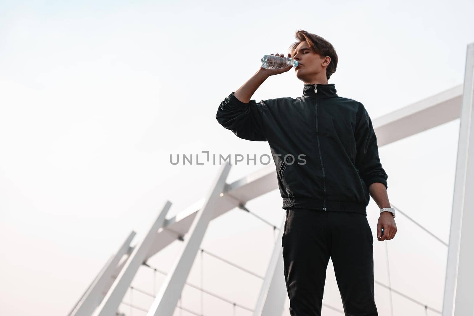 Young stortsman is drinking water near the stadium. He wears black sport clothes