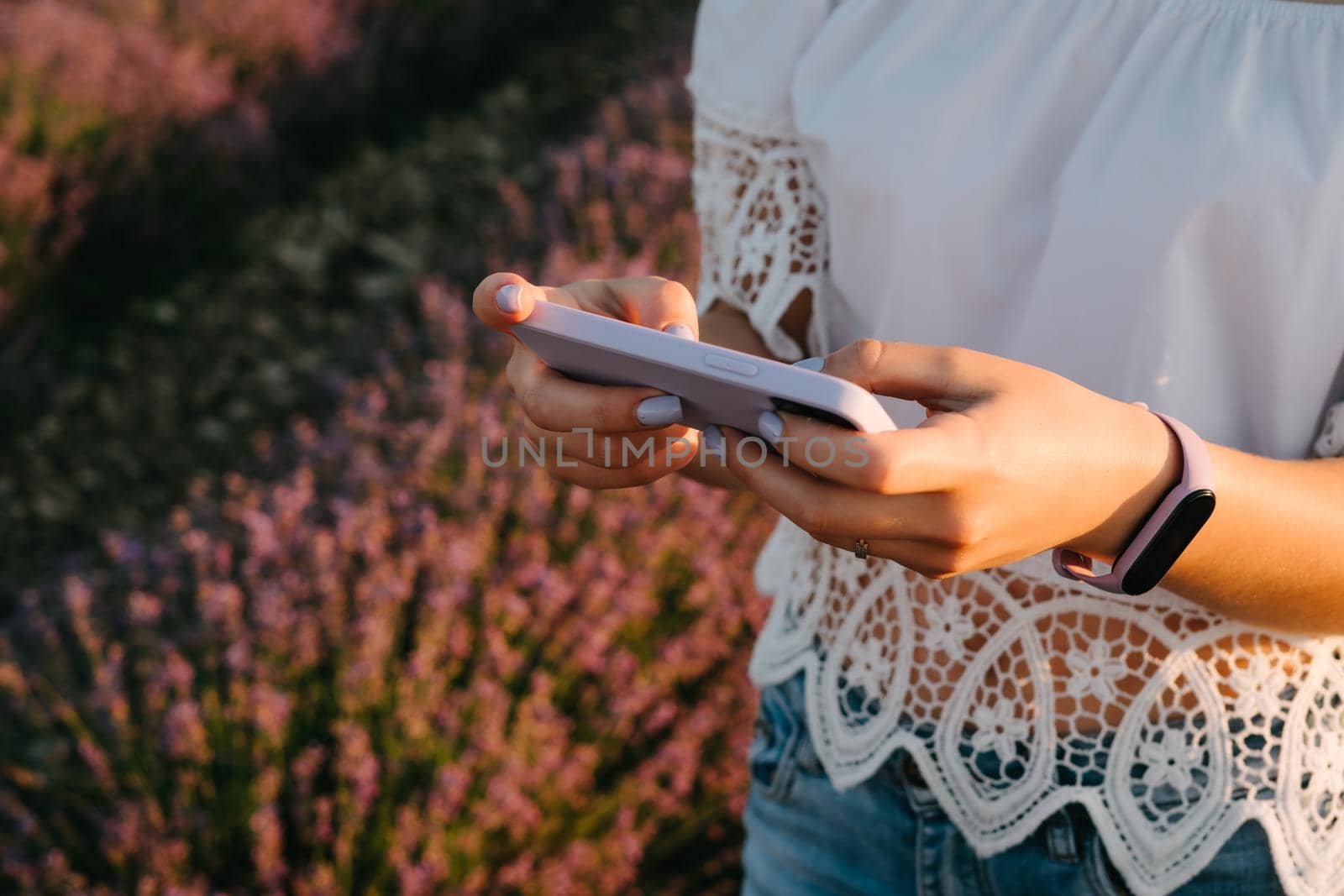 A girl in a white cotton top with lace holds a smartphone in a purple case. Beautiful lavender field. Modern technologies. Fitness bracelet with purple silicone strap. Waterproof smart watch.