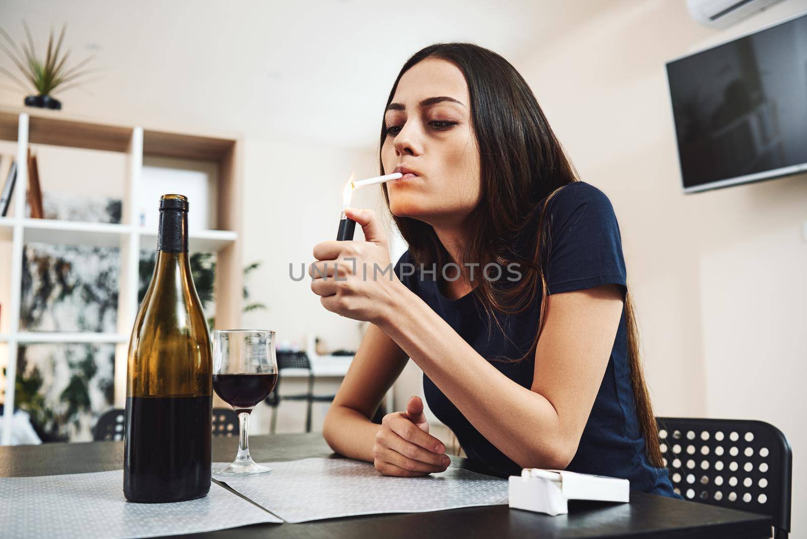 Portrait of young woman in despair, smoking cigarette while sitting at table with glass of alcohol at home. Female alcoholism concept. Protest in the treatment of alcohol addiction.