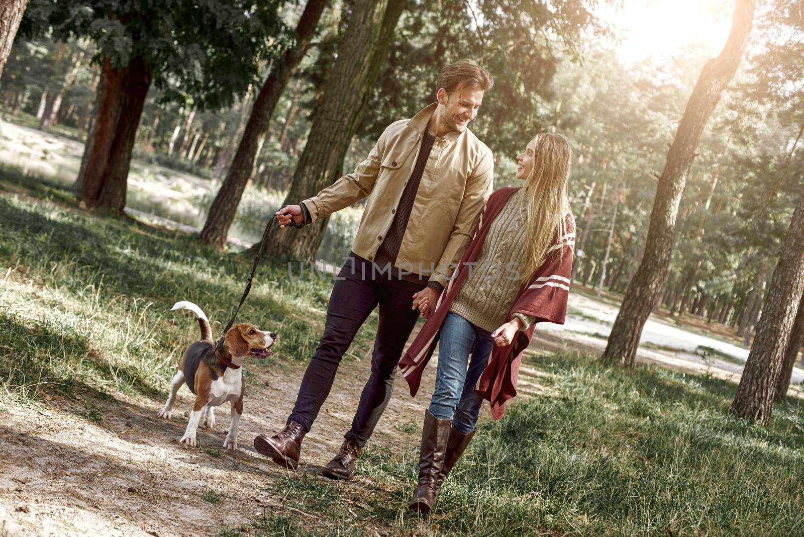 Young family couple are walking in woods with their dog. Stylish man is looking at his girlfriend while holding her hand. Bright sun is shining throw the trees, outumn season.