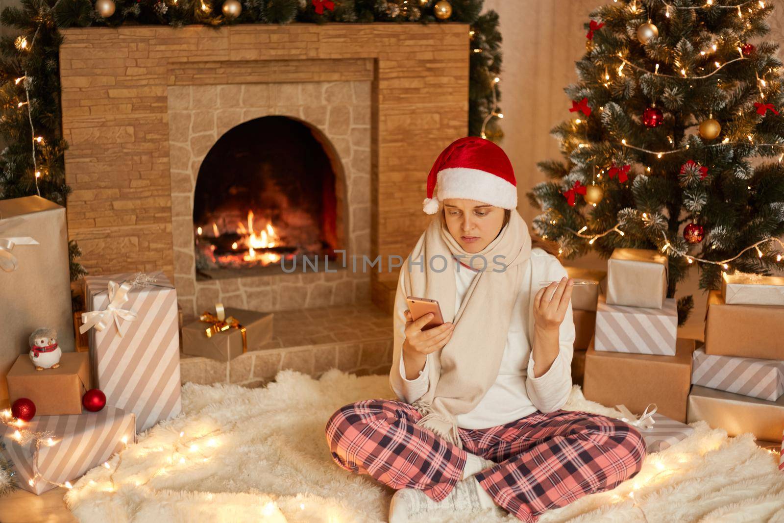 Sad sick woman in santa hat and mask using cellphone for video call and showing thermometer to camera, spending holidays at home because of pandemic, sitting on floor with crossed legs on floor.