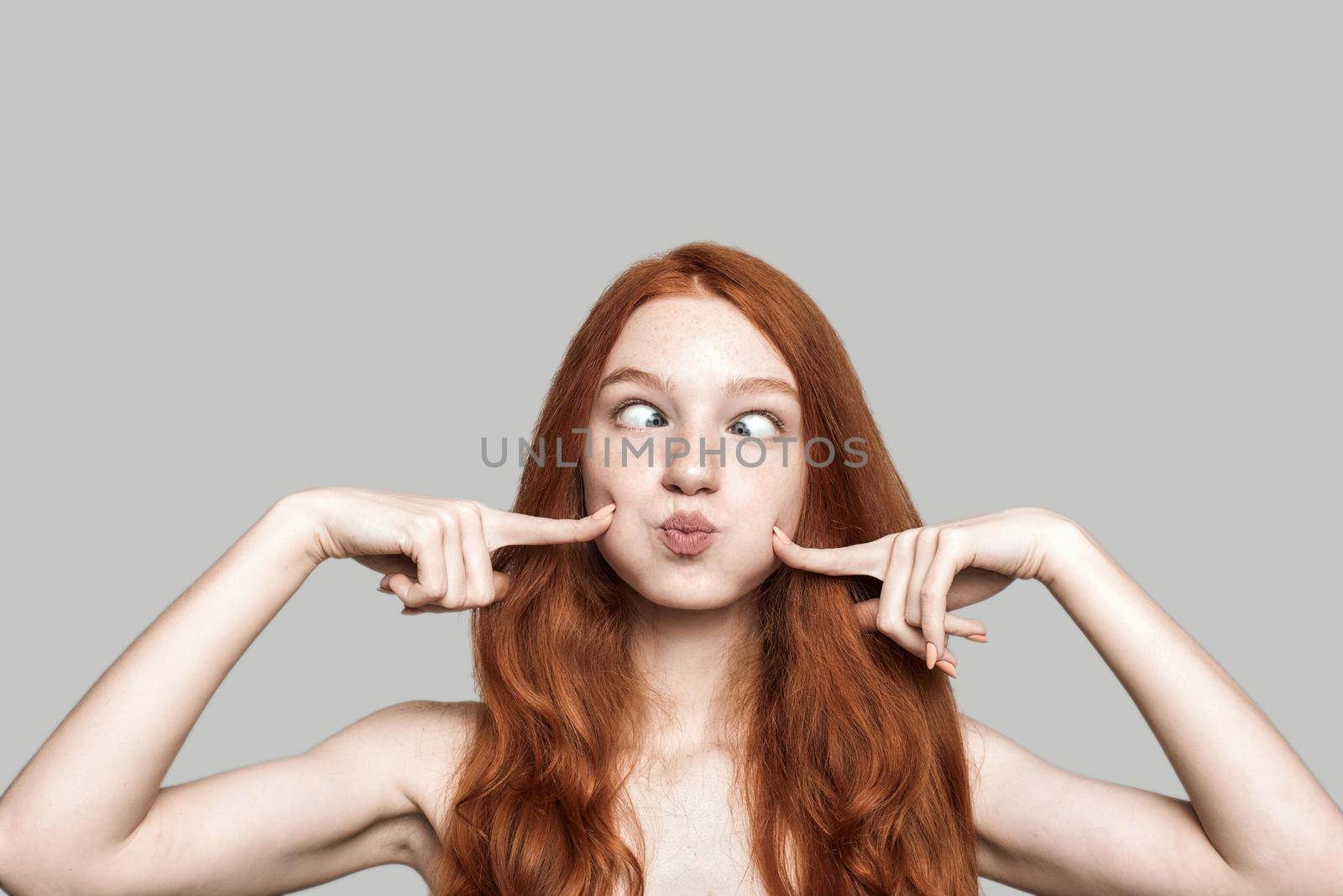 Studio shot of happy young redhead woman making crazy face and grimacing while standing against grey background