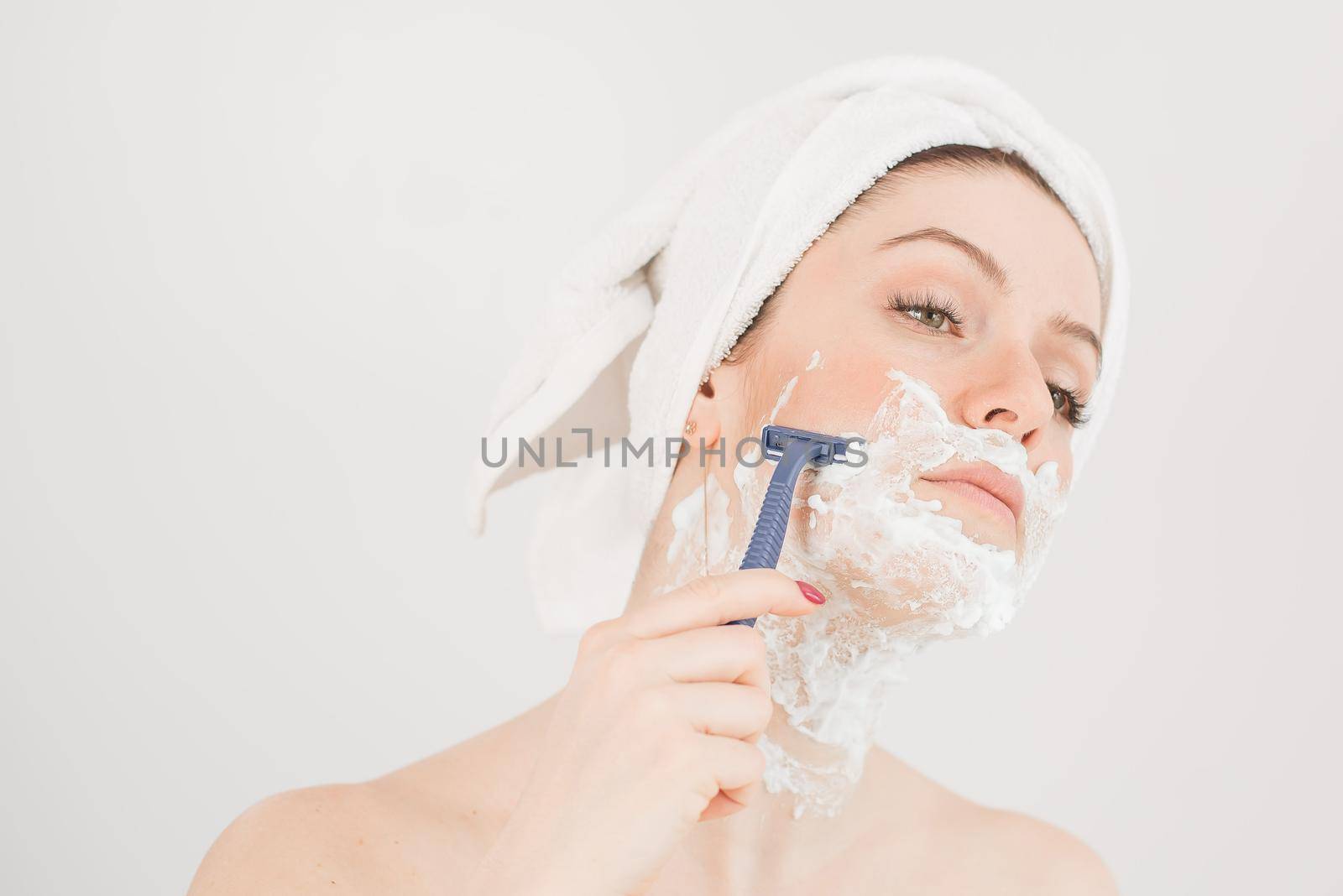 Cheerful caucasian woman with a towel on her head and shaving foam on her face holds a razor on a white background by mrwed54