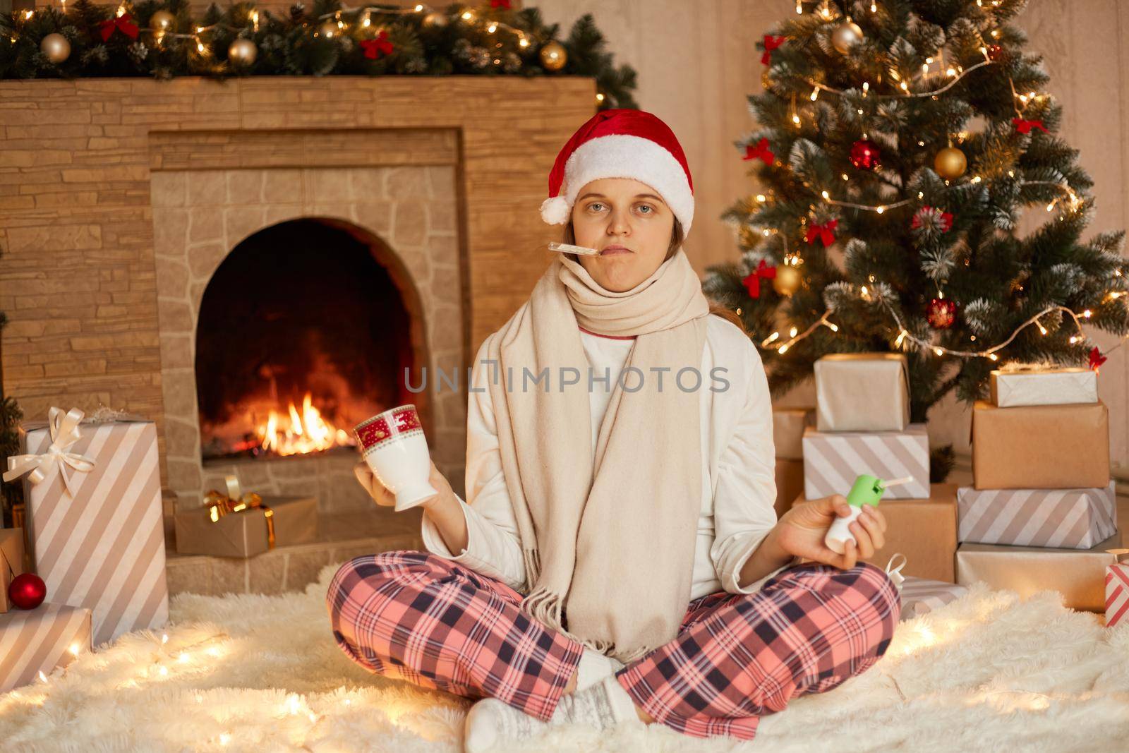 Sad sick girl with thermometer in mouth on Christmas holiday drinking tea and holding sore throat spray, looking at camera with unwell facial expression, wearing casual clothing, wrapper scarf and hat