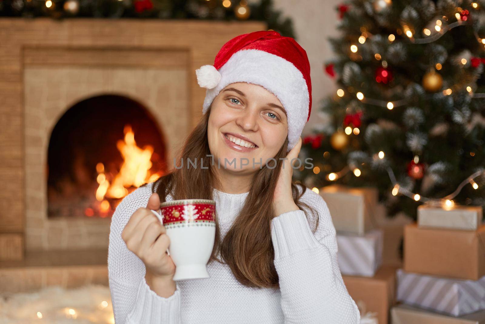 Woman with cup in hands sitting in living room near fireplace, looking at camera with toothy smile, enjoying hot beverage in cozy atmosphere, girl posing near Christmas tree and present boxes.