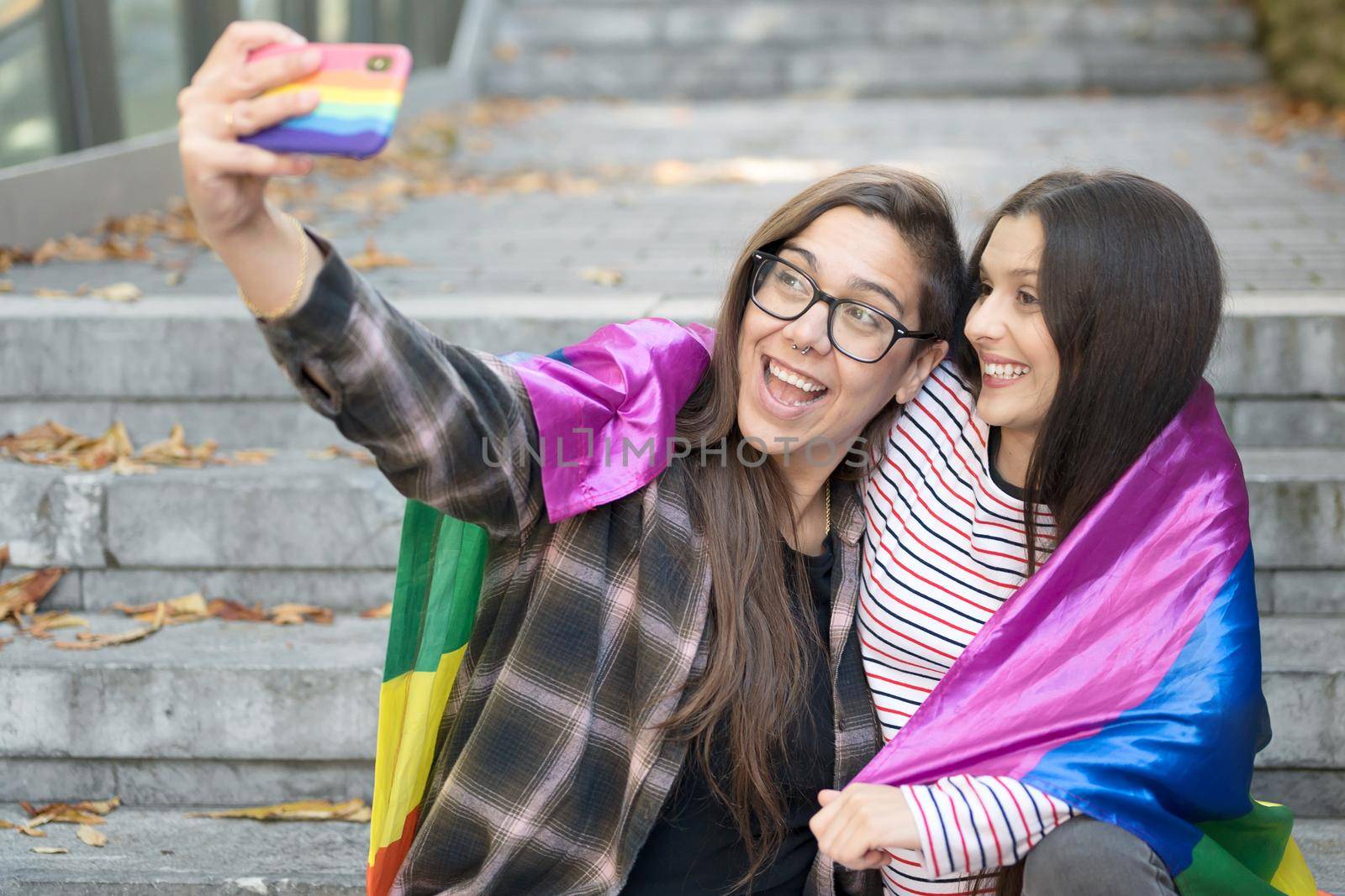 Cheerful happy lesbian couple holding lgbt flag taking a selfie outdoors. by HERRAEZ