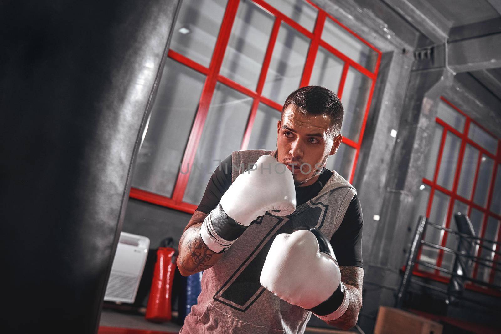 One second before. Focused tattooed athlete in sports clothing boxing on heavy punch bag while exercising in the boxing gym by friendsstock