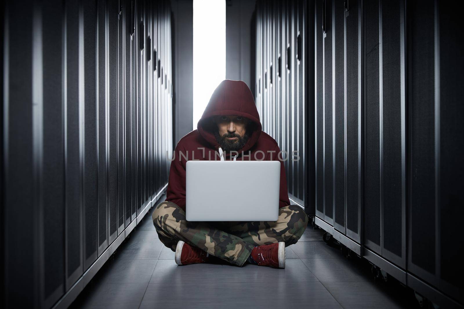 The hacker in a hood sits with the computer among servers, the burglar, face covered with a hood. Storage of data, cloudy services, system breaking, attack of hackers by vladimirdrozdin