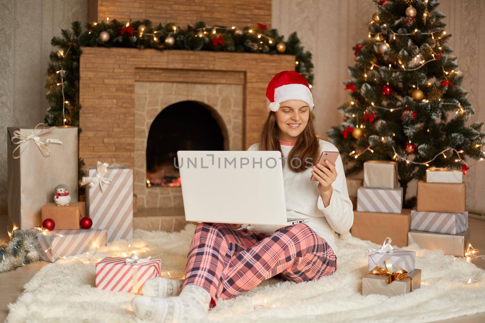 Happy beautiful young woman with laptop on knees sitting near Christmas tree and fireplace on floor, holding phone in hands, sending message with congratulating, looks glad. by sementsovalesia