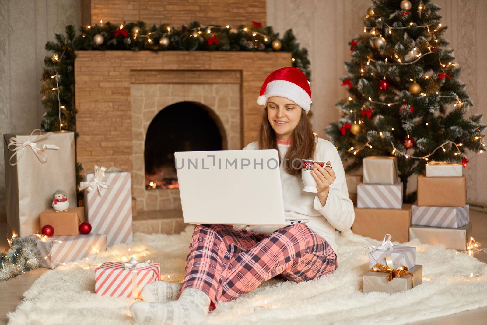 Merry Christmas and happy new year, smiling woman meeting together with somebody online via video calling on laptop, drinking coffee or tea while sitting on floor near fireplace.