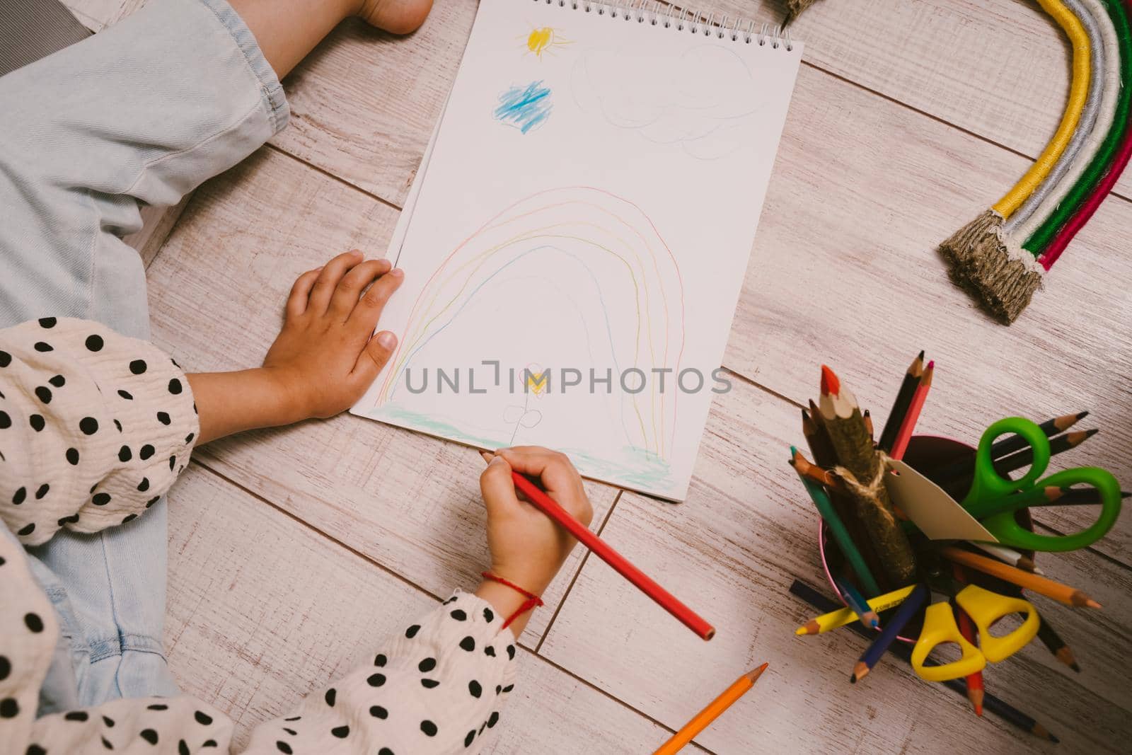 A little girl sits on the floor and draws in a sketchbook with colored pencils. The child draws flowers and rainbows. Pencils in the hands of the girl.