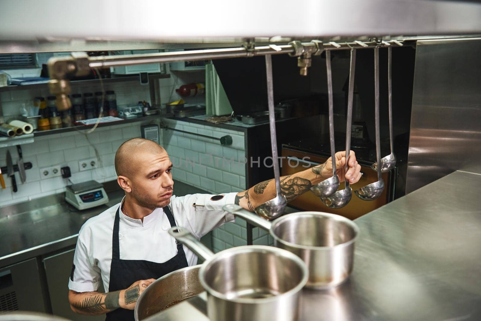 Cooking soup. Young male chef in apron choosing a ladle for his famous soup while standing in a restaurant kitchen by friendsstock