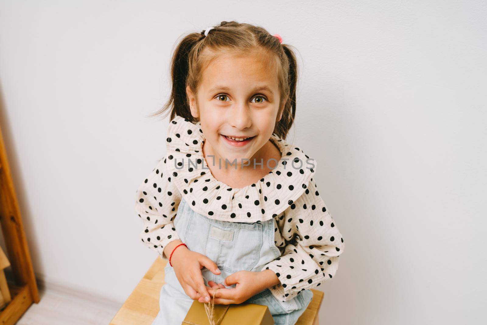 Funny girl sits on a wooden stool with a gift on her knees. A little girl in a polka dot print shirt. The child smiles.