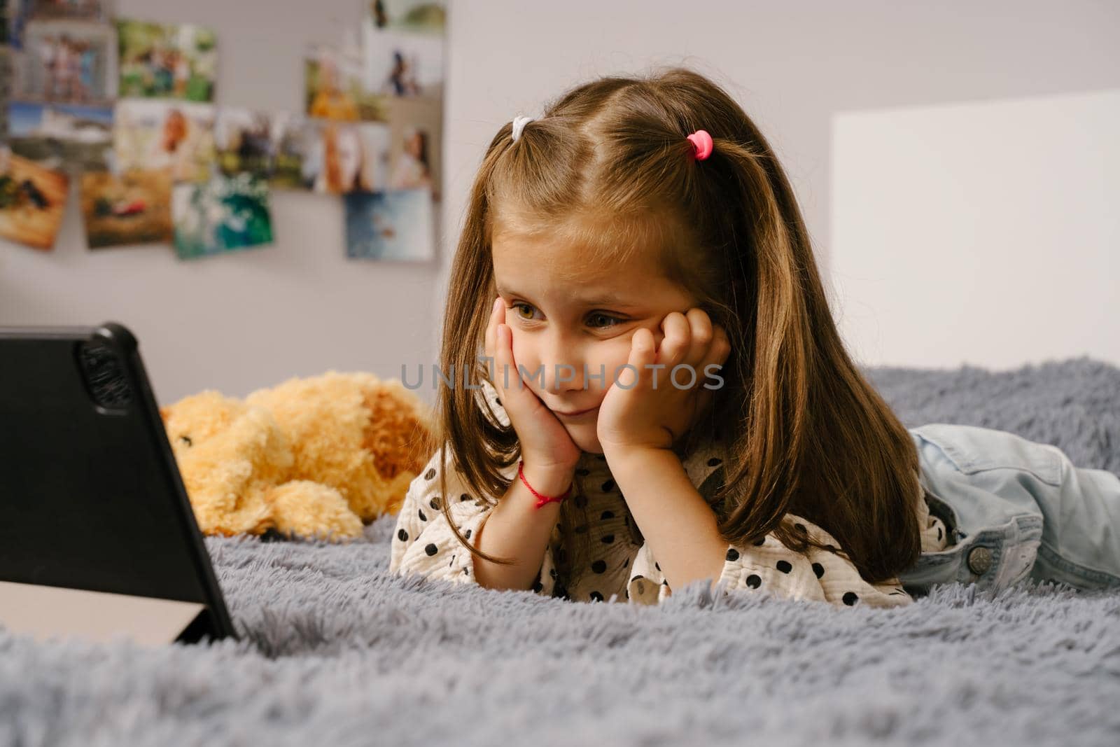 The child lies on the bed and watches cartoons on the tablet. A little girl in a polka dot print shirt. The child smiles. A little girl is talking very emotionally over a video link.