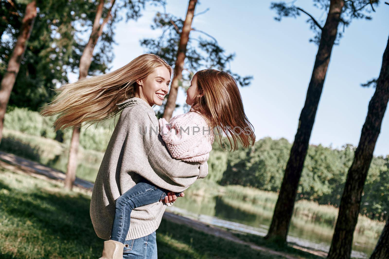 A little curly girl in sweater is hugging her mother in woods. Cold season, bright sun is seen through the trees. She is wearing pink sweater and young woman is in grey sweater