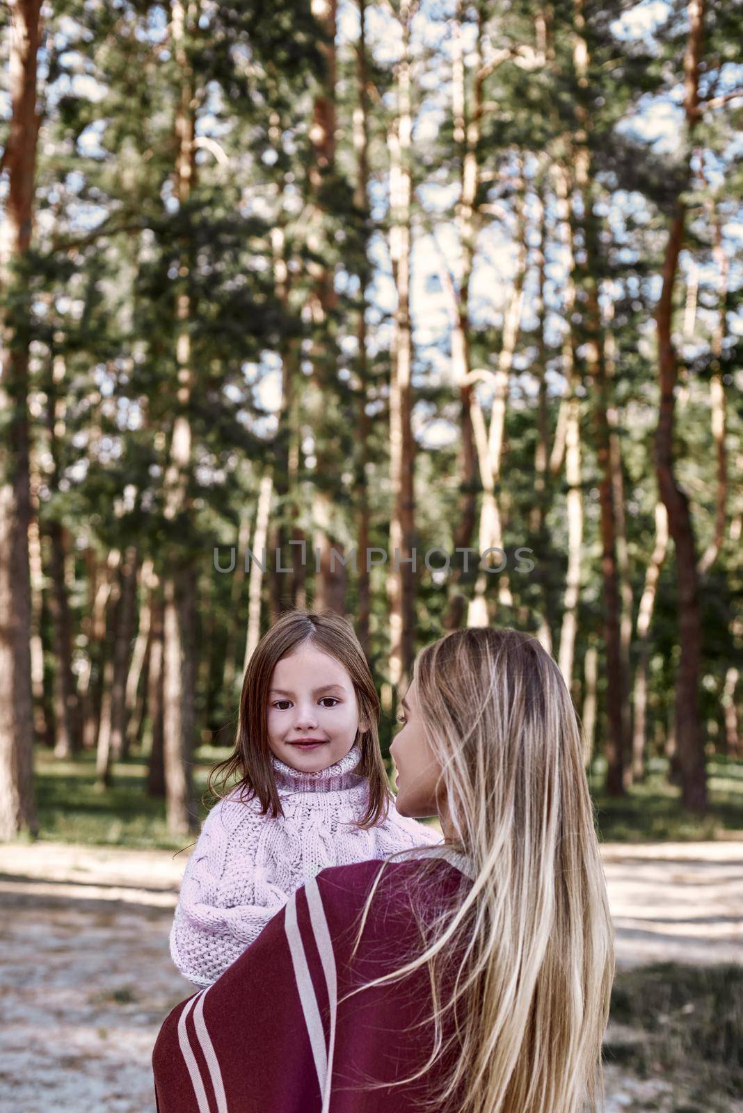 A little curly girl in sweater is hugging her mother in woods near lake. She is wearing pink sweater and young woman is in red knitted coat.