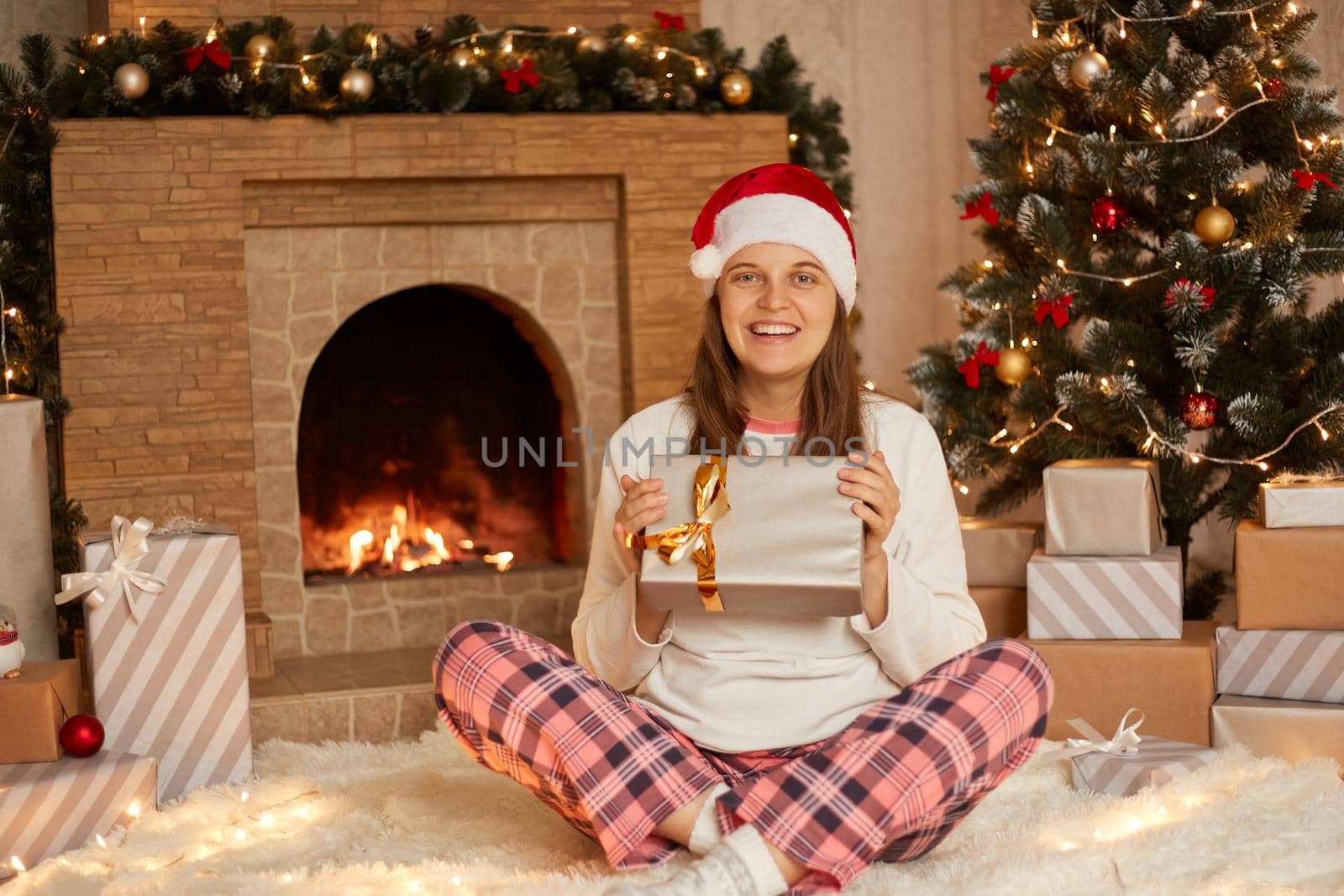 Happy smiling woman in casual clothing and santa claus hat sits on floor on warm soft carpet with crossed legs and holds gift box, looks at camera, being satisfied, poses near fireplace and fir tree.