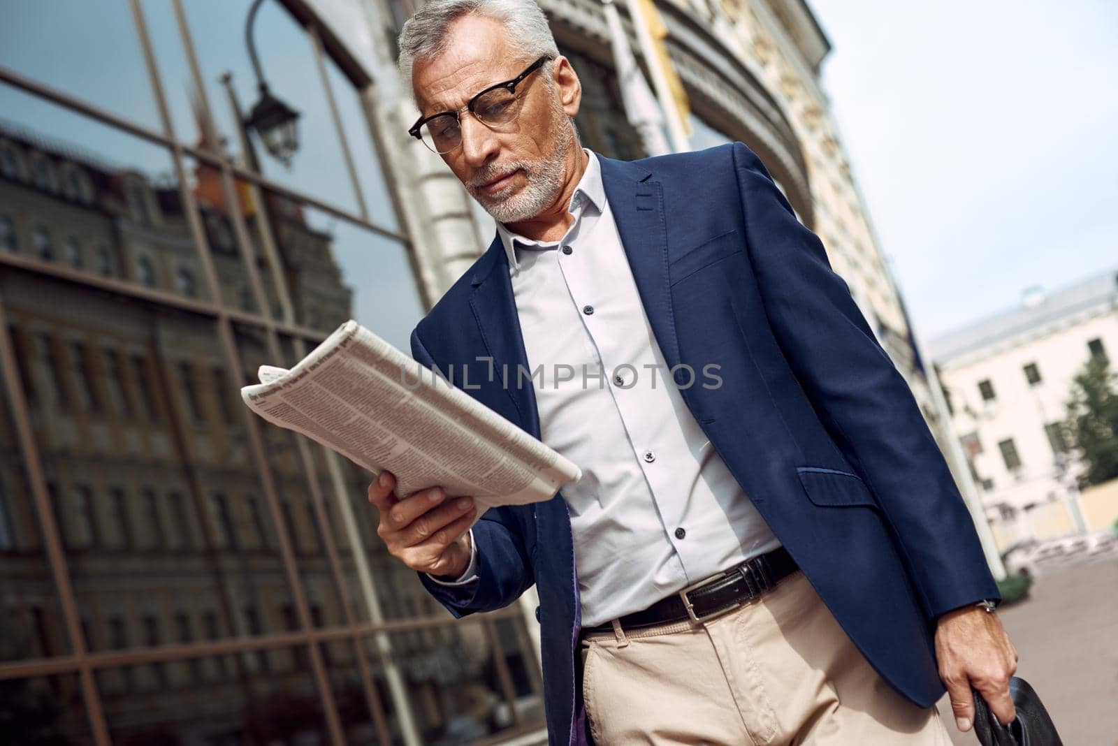 Reading the latest news. Confident senior man in casual suit reading newspaper while standing outdoors with cityscape in the background by friendsstock