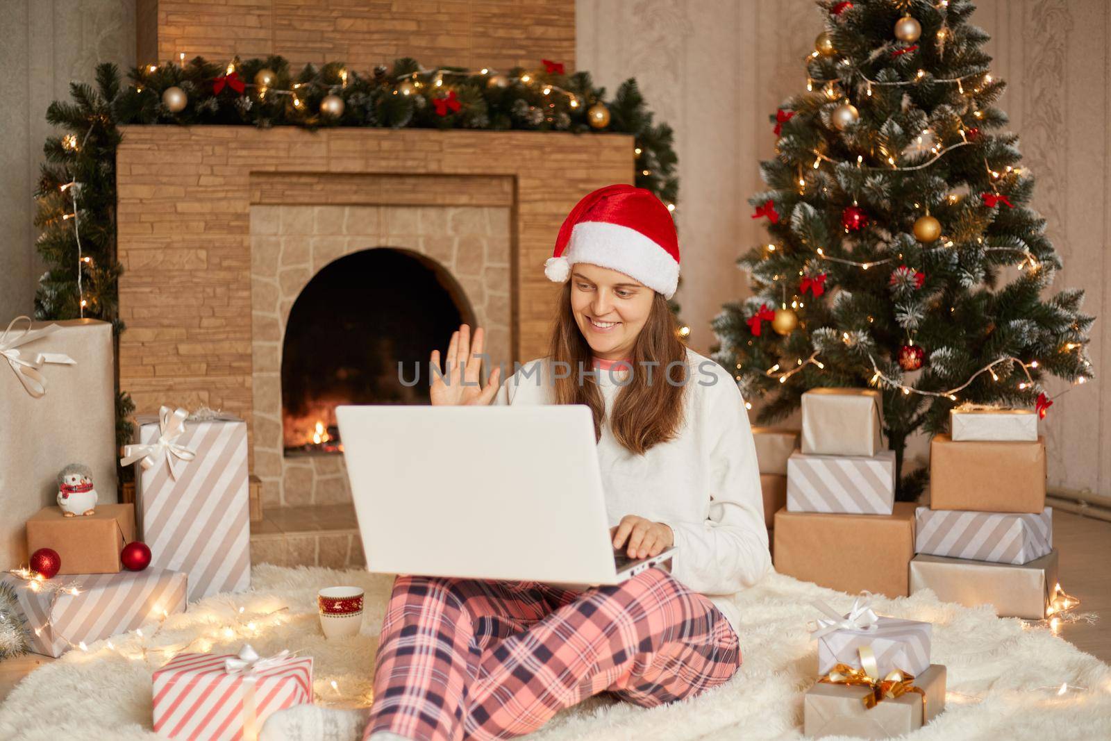 Beautiful young woman sitting on floor with laptop on knees, having video call, greeting somebody and waving hand, wearing checkered pants, white shirt and santa claus hat.