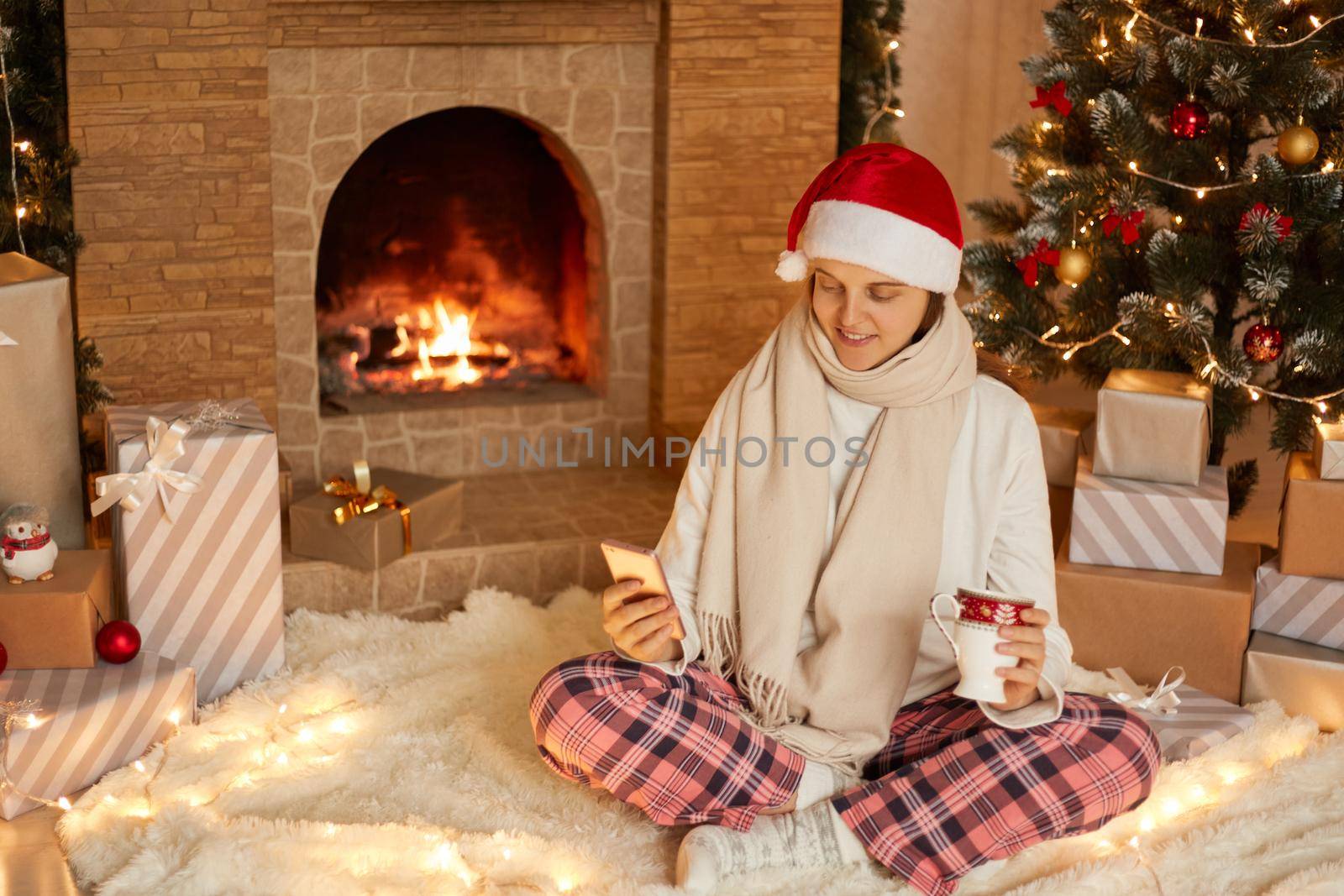 Photo of positive lady sitting with cellphone and holding beverage mug in house indoors with christmas x-mas ornament, wearing santa hat, scarf and checkered pants, having video call.