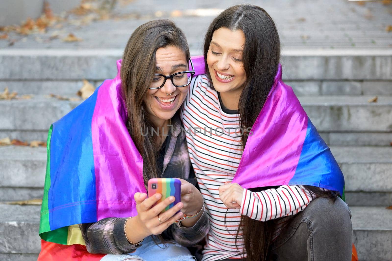 Beautiful lesbian couple with rainbow flag using a mobile phone in the street. by HERRAEZ
