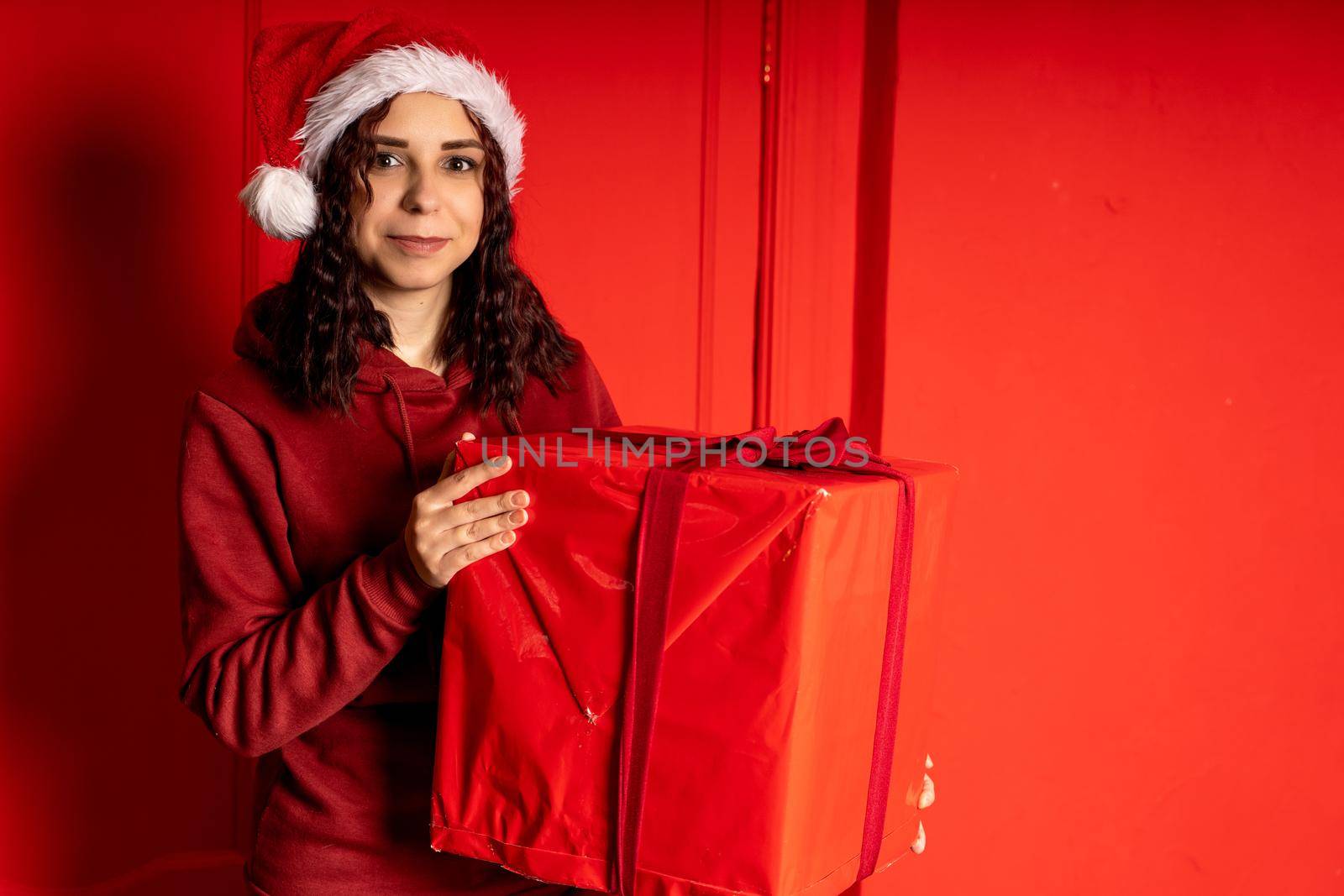 Young woman in Santa Claus hat holds big gift, standing near red wall. Happy female in Christmas hat with box of gift. Concept of holidays, presents and good mood. by epidemiks