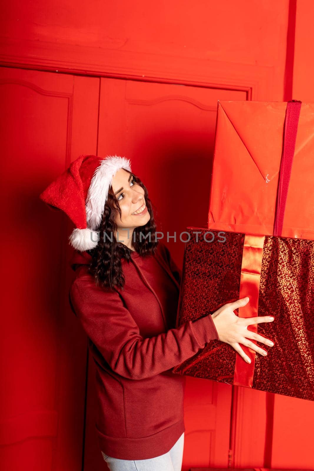 Young woman in Santa Claus hat holds big gifts, standing near red wall. Happy female in Christmas hat with boxes of gift. Concept of holidays, presents and good mood. by epidemiks