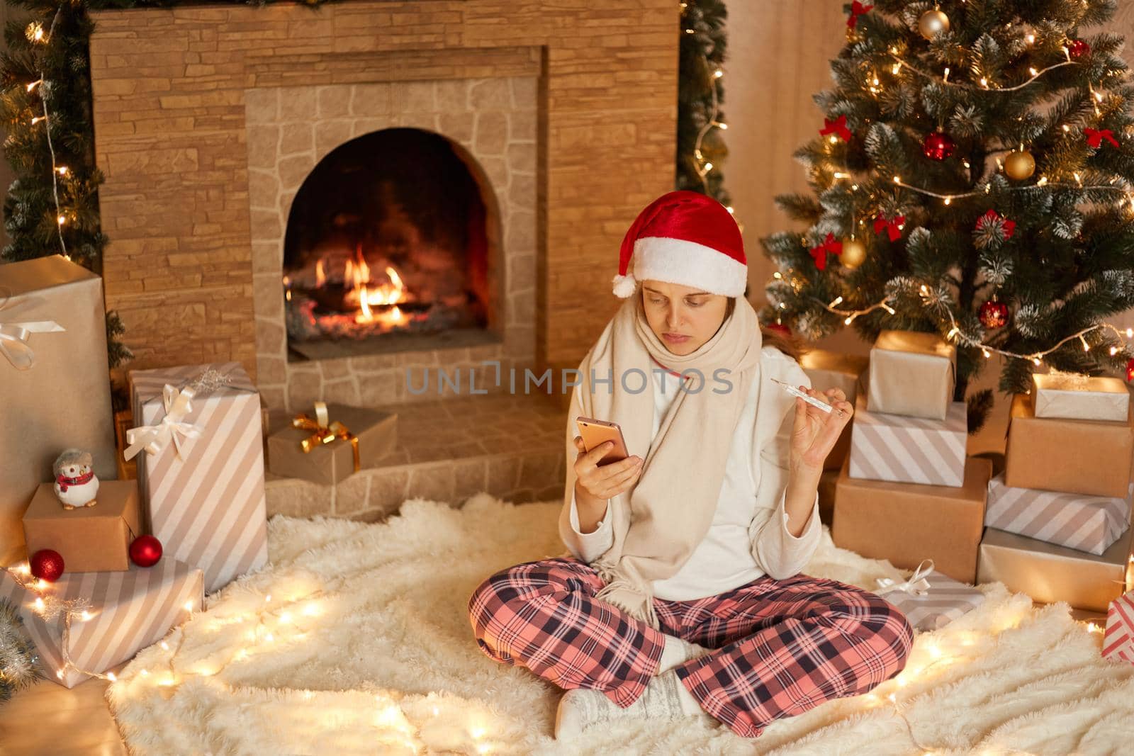 Young woman wearing santa hat, scarf and checkered pants, take video calls with somebody and showing thermometer to camera, telling about her high temperature, posing in festive living room.