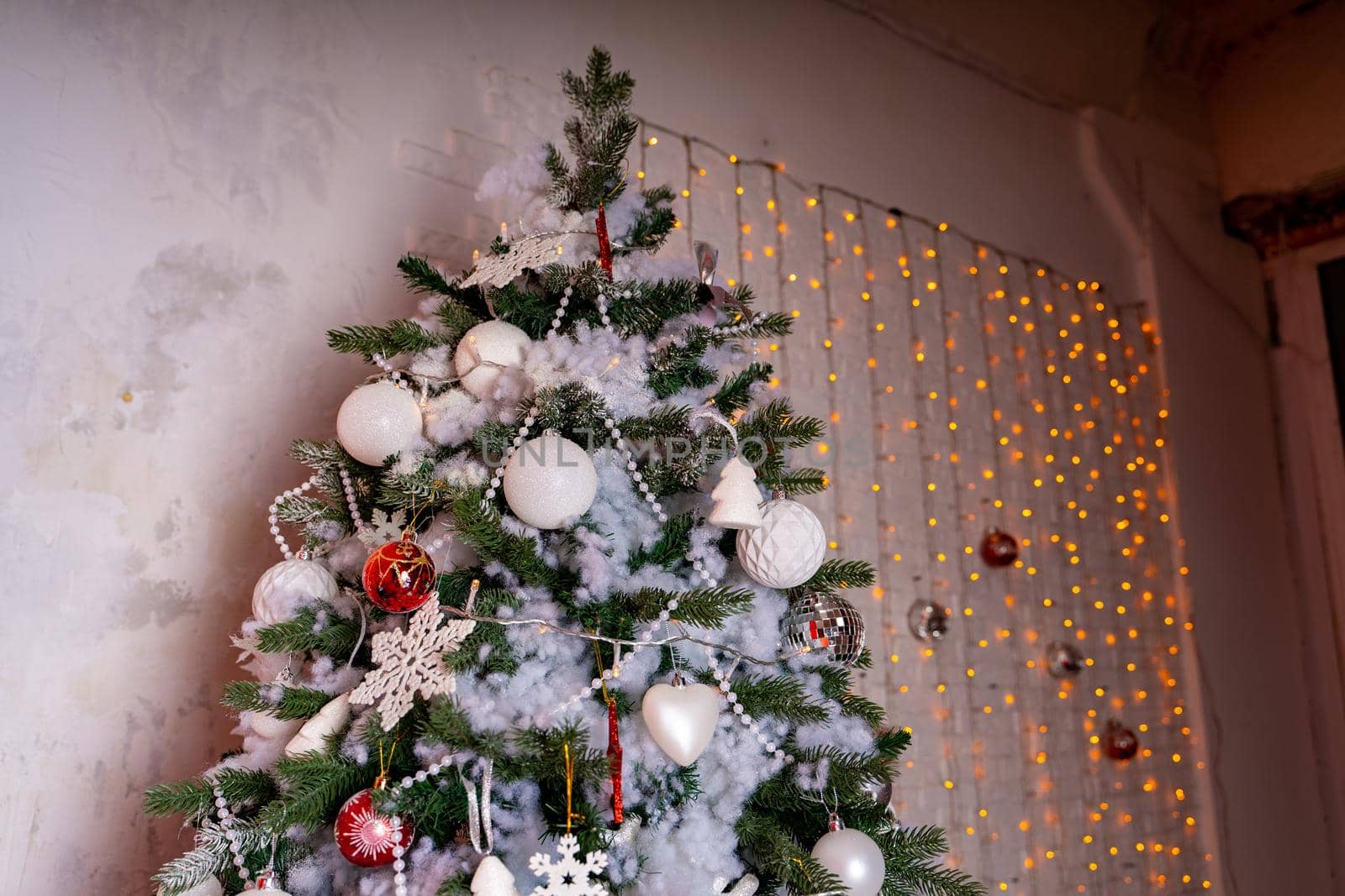 Decorated Christmas tree in the room. Coniferous tree with white and red baubles located near the white wall during the celebration of the holiday.