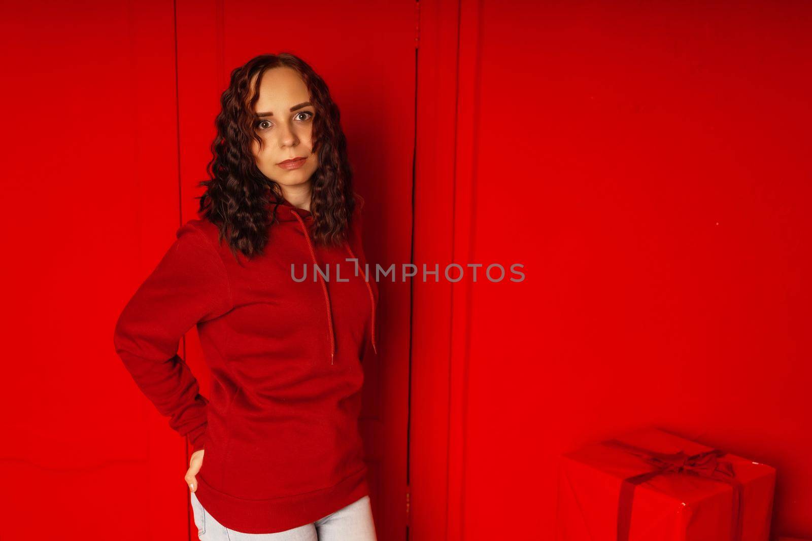 Young woman in hoodie on red background. Curly brunette poses near red wall. by epidemiks