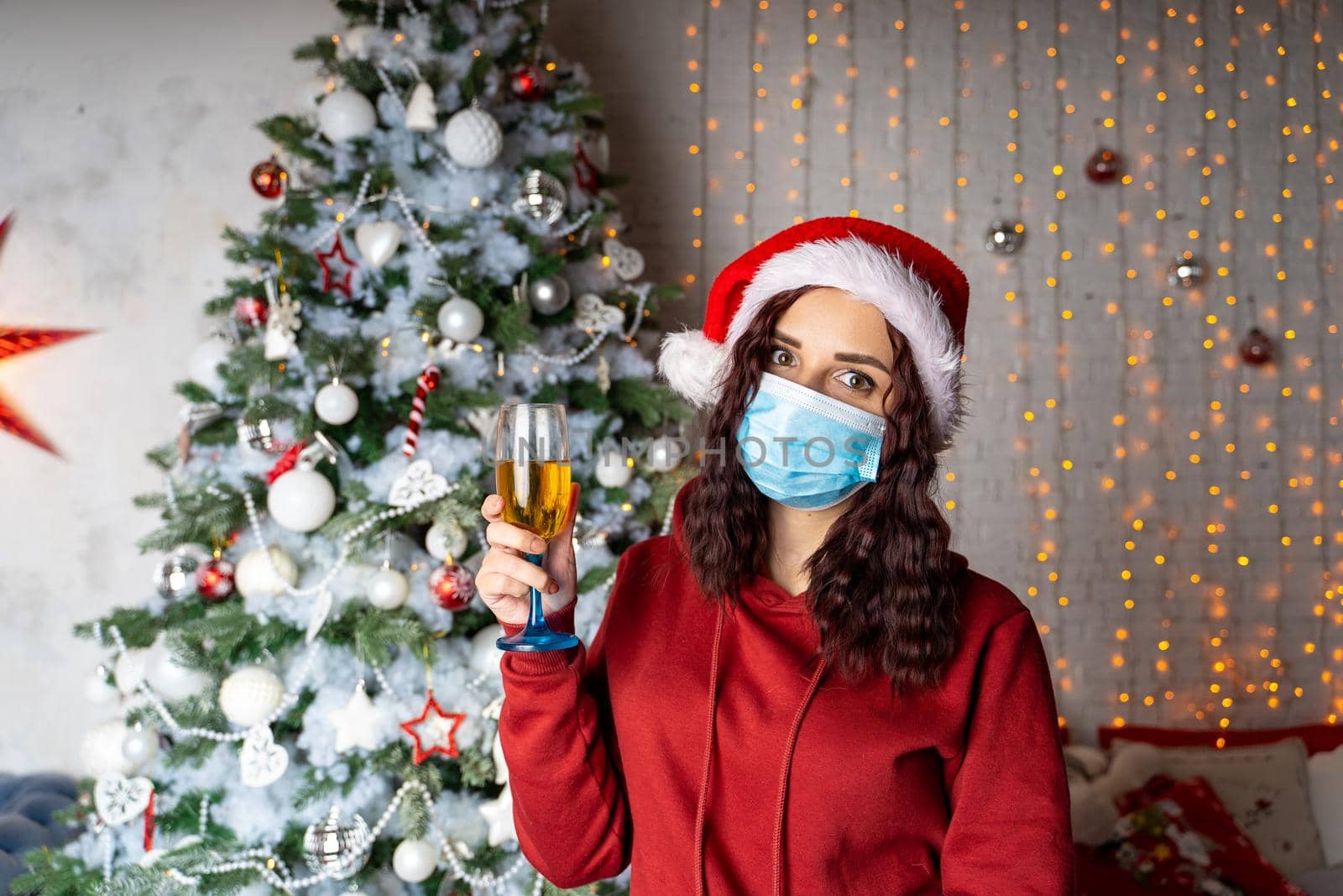 Young woman in medical mask and santa hat with glass of champagne on background of Christmas tree. Charming brunette in protective mask celebrating of safe Christmas during coronavirus pandemic