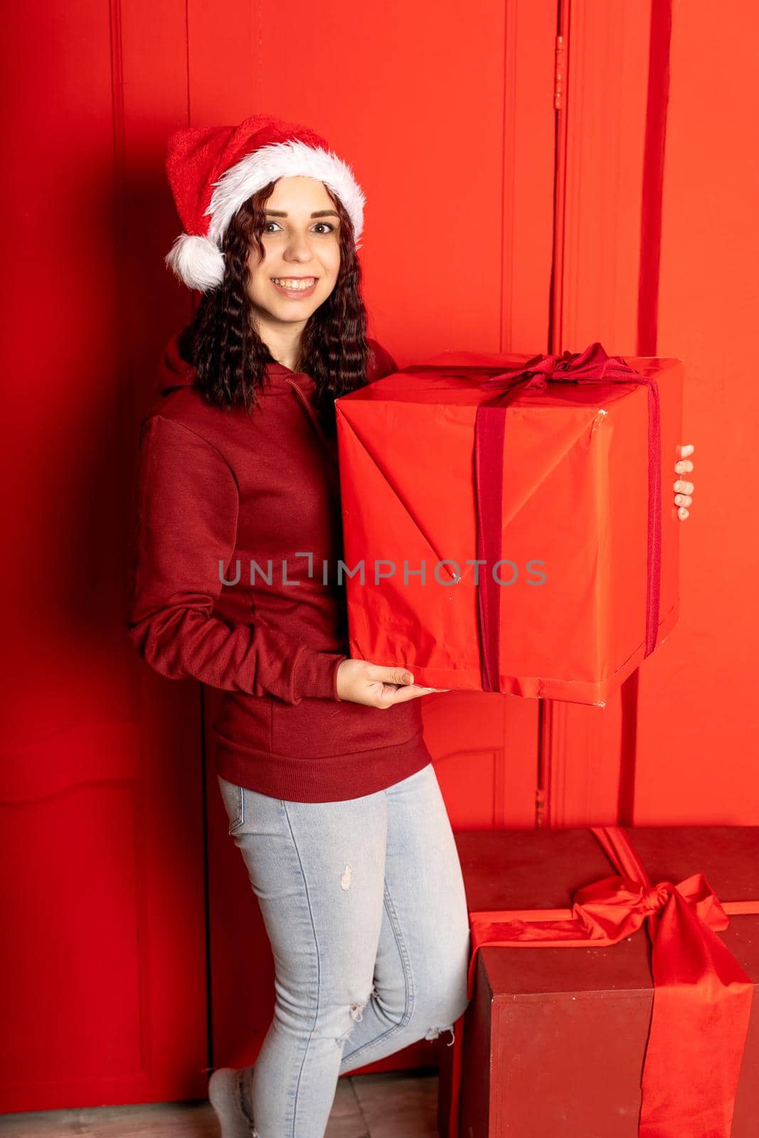 Young woman in Santa Claus hat holds big gift, standing near red wall. Happy female in Christmas hat with box of gift. Concept of holidays, presents and good mood. by epidemiks