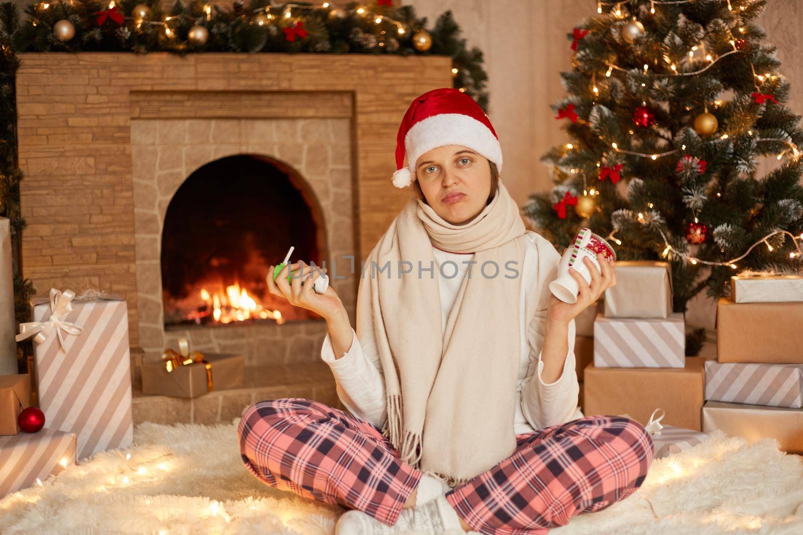 Young sick girl in santa hat and checkered pants, holding spray and cup of warming drink in hands, looks at camera with sad facial expression, being in during Christmas holidays.