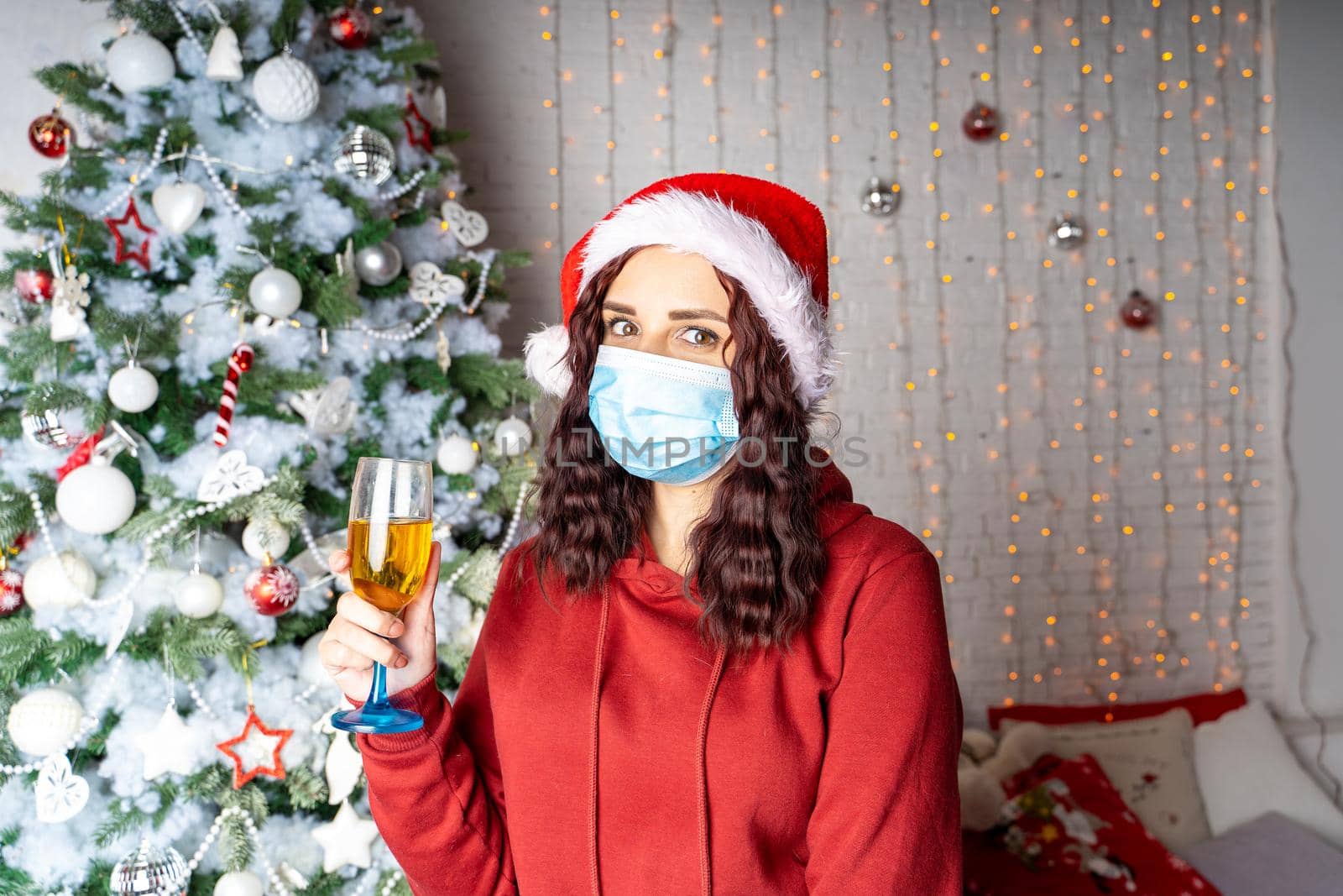 Young woman in medical mask and santa hat with glass of champagne on background of Christmas tree. Charming brunette in protective mask celebrating of safe Christmas during coronavirus pandemic. by epidemiks