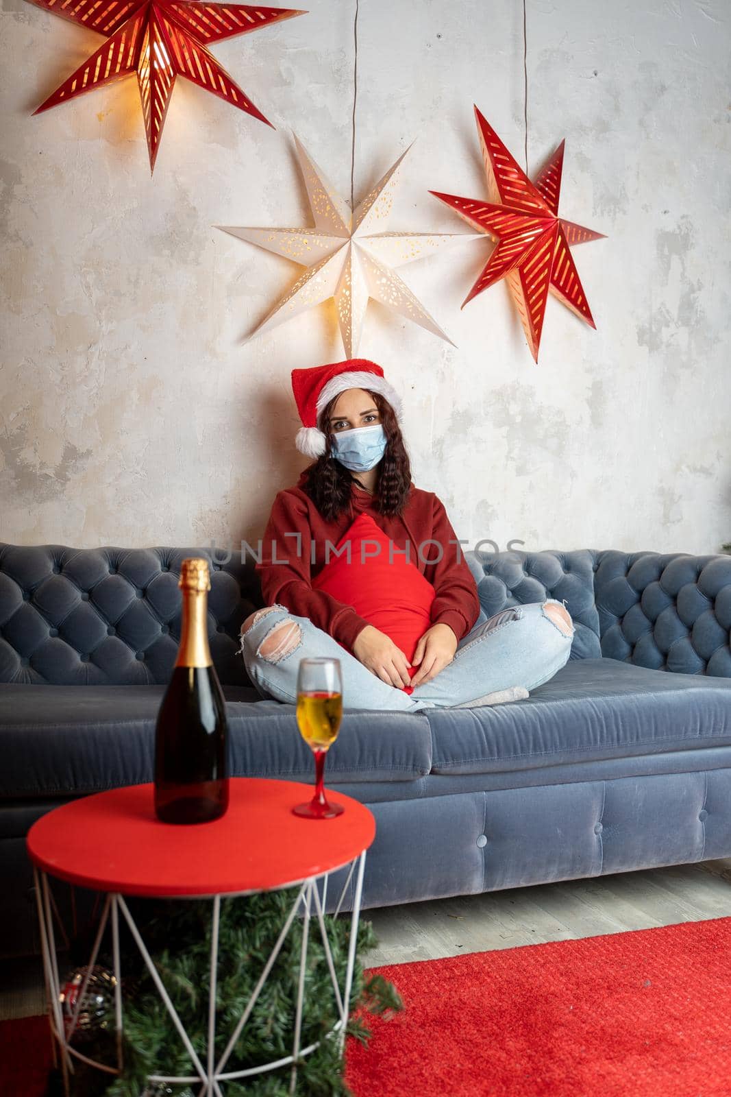 Young woman in medical mask and Santa Claus hat sitting on sofa in room. Alone charming brunette in protective mask celebrating of safe Christmas during coronavirus pandemic. by epidemiks