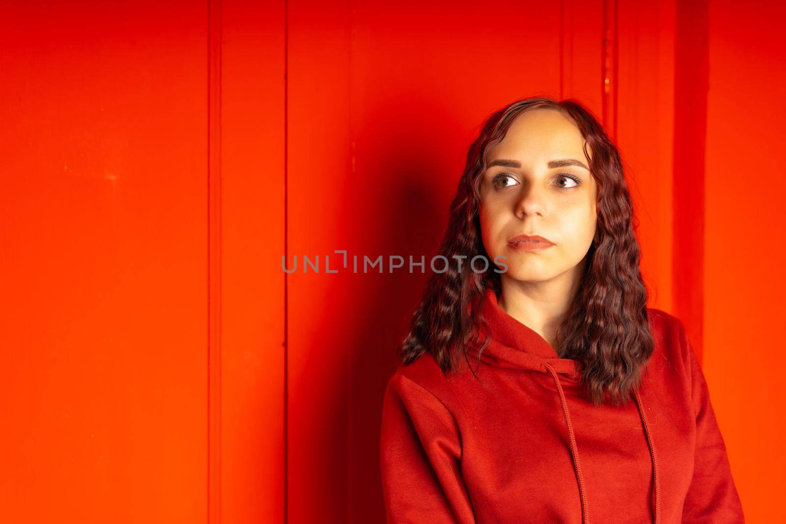 Young woman in hoodie on red background. Curly brunette poses near red wall. by epidemiks